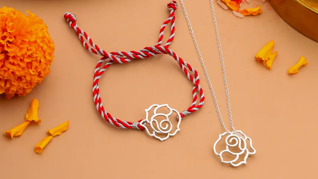 A silver rose motif Rakhi on a red and silver thread, displayed alongside a matching silver pendant on a chain and traditional puja items, illustrating a versatile Rakhi designed to be worn as a keepsake necklace after Raksha Bandhan.